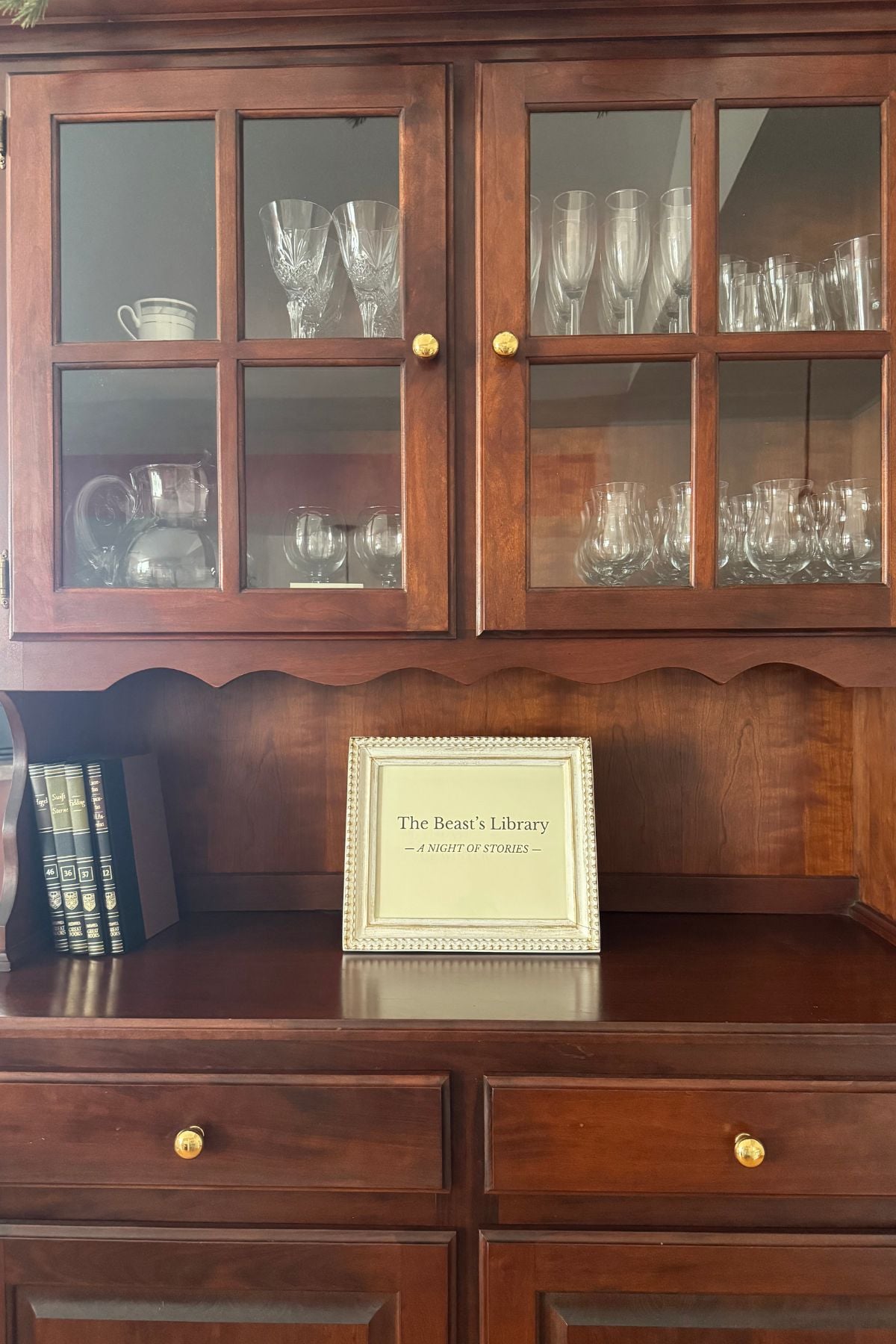 A framed sign: The Beast's Library - a night of stories - in a china cabinet with four books leaning against the side of the cabinet