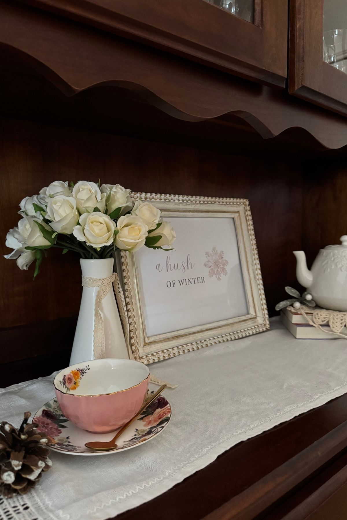 Winter vignette on China cabinet buffet: soft white roses in a white vase, pink tea cup, a framed sign: a hush of winter, a white teapot.