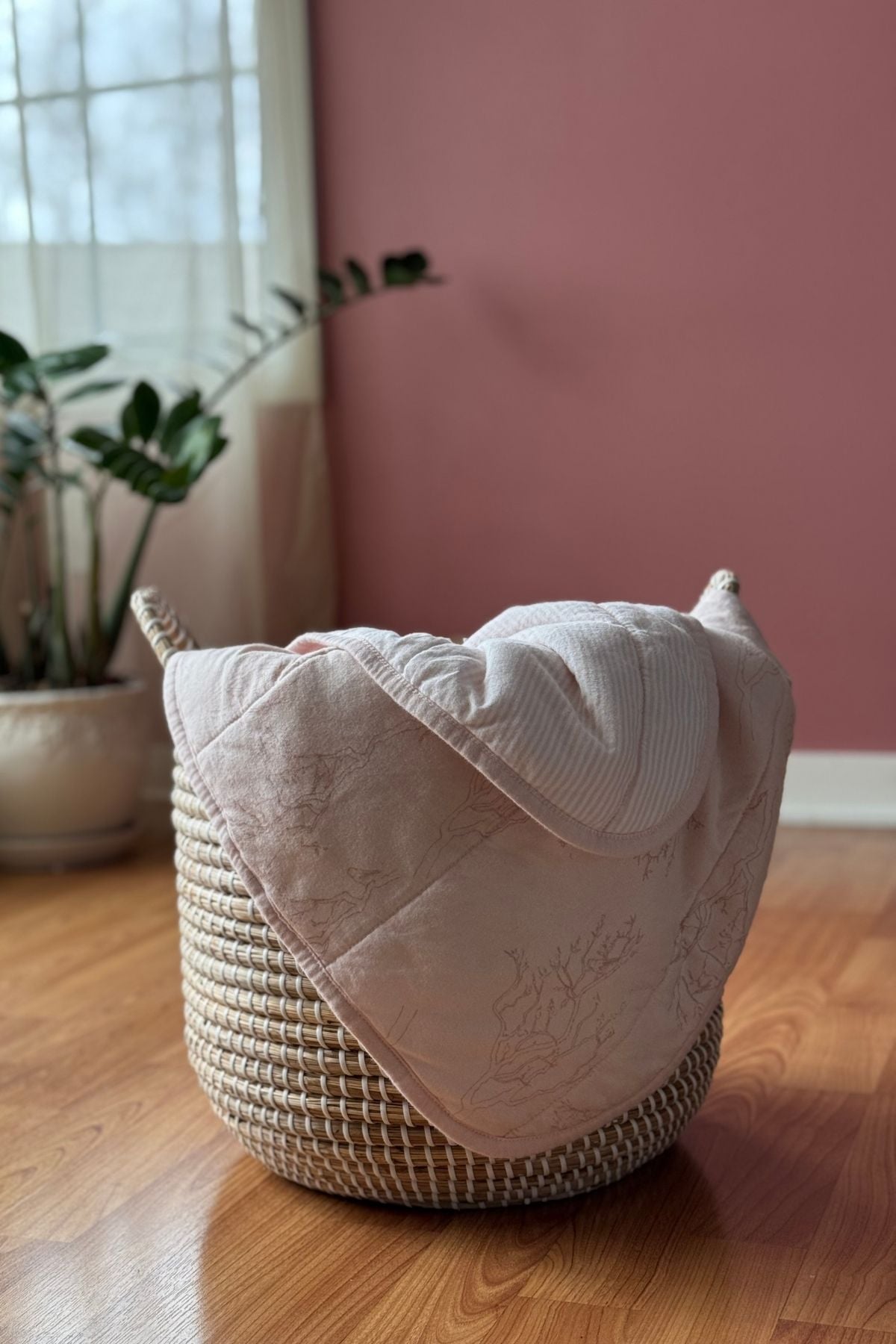 A blush pink reversible organic cotton jersey baby blanket in a woven basket in the living room.