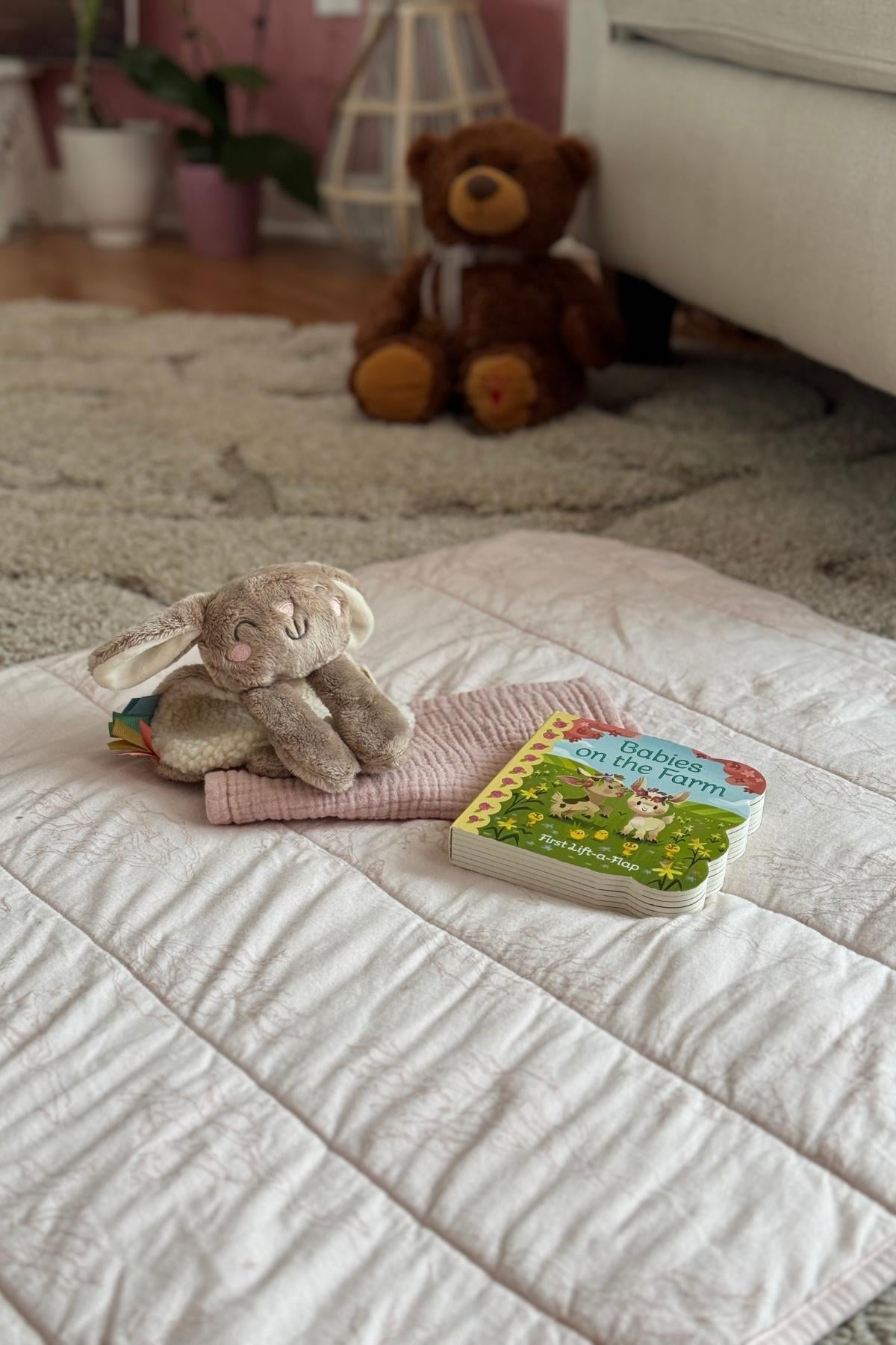 A blush pink organic cotton baby blanket on the living room floor with a book, burp cloth, and a little bunny