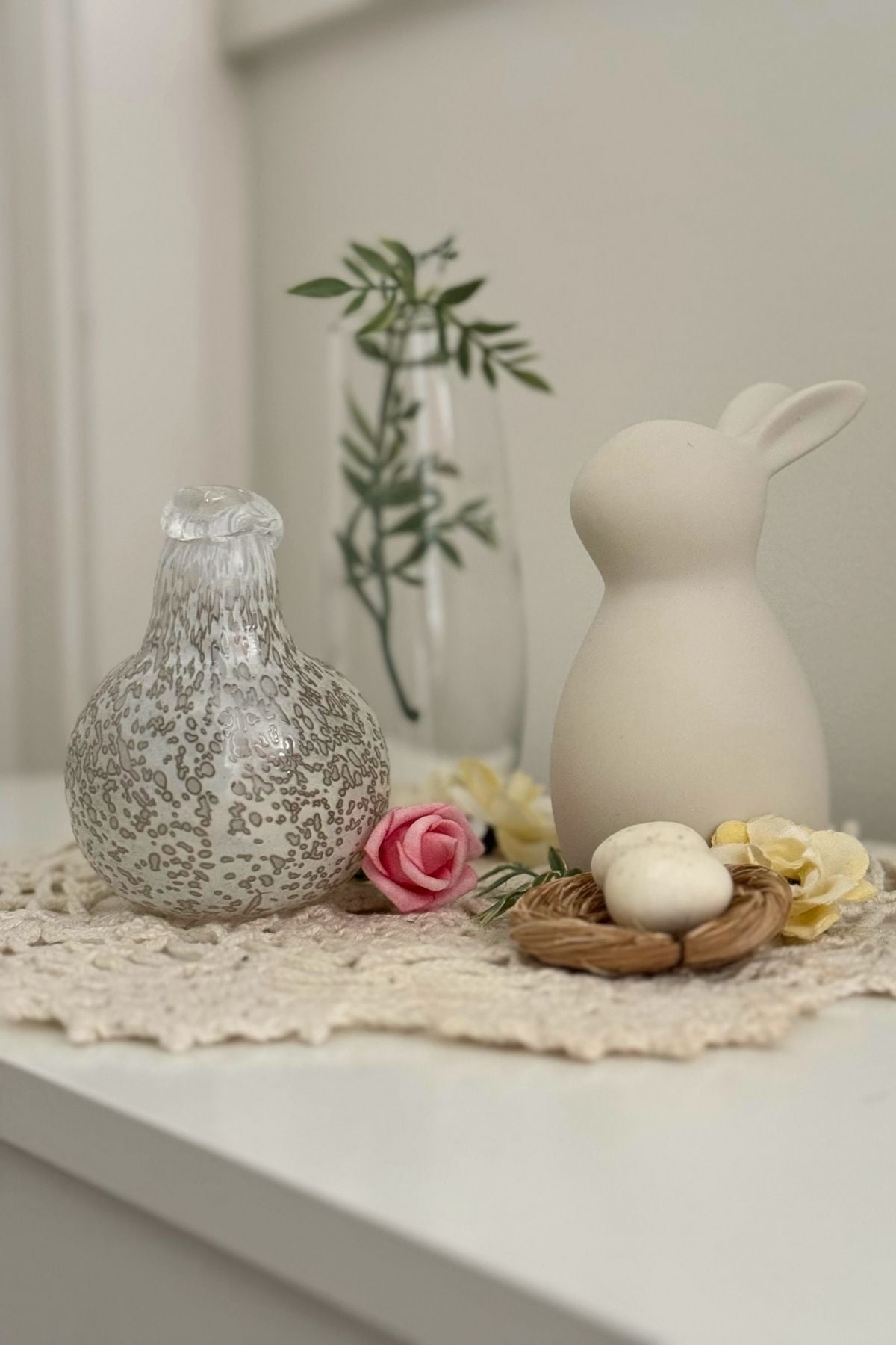 grandmacore spring decor for hallway Spring entryway table with a crocheted runner, glass vase with greenery, bunny figurine, glass bird, and a small nest with speckled eggs in soft neutral tones