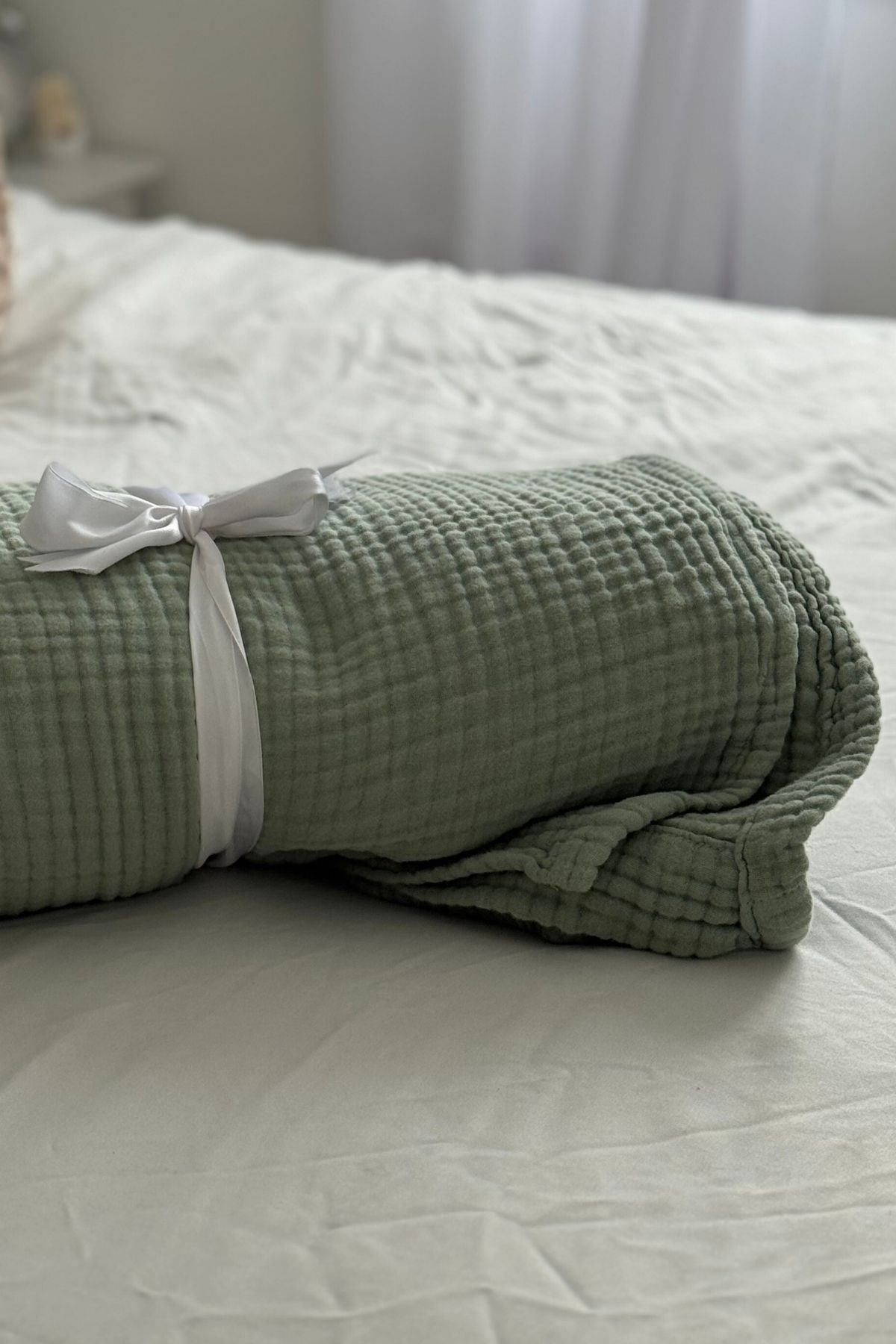 A sage green twin-size muslin blanket rolled up and wrapped with a white ribbon.