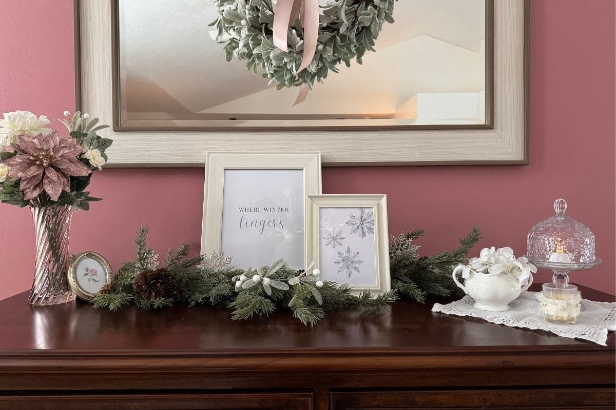 A dark wood sideboard with two white framed signs, evergreen branches, and blush and white decorations.