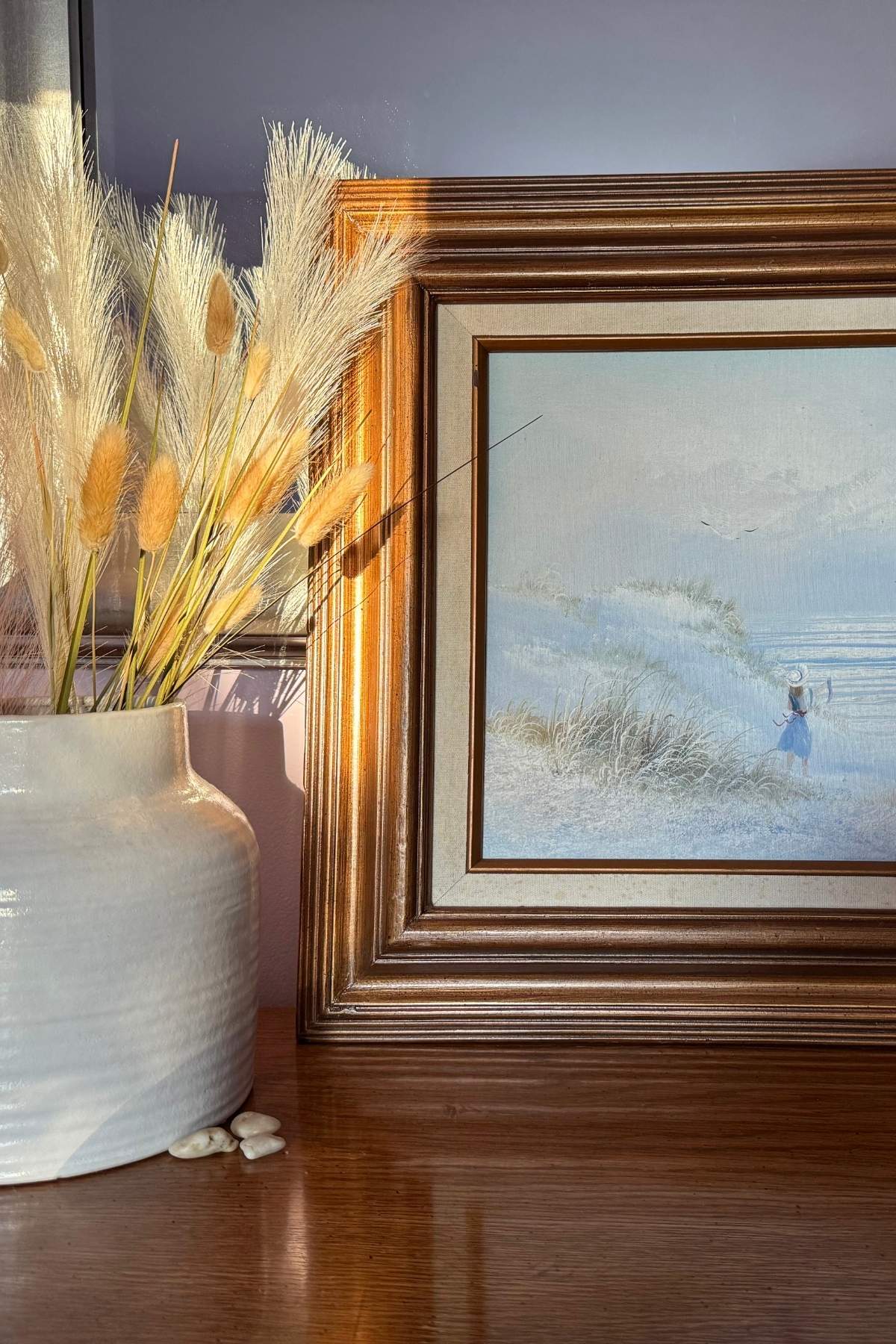 Simple decor for master bedroom. A white vase holding weathered grass next to painting of a quiet shoreline.