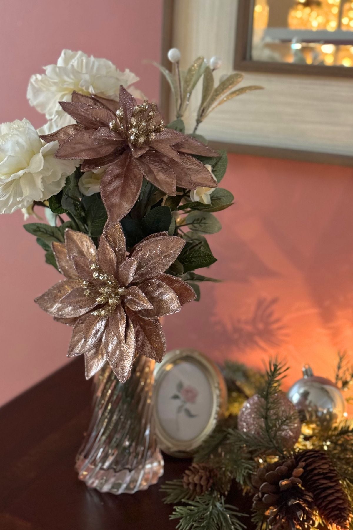 Christmas florals blush poinsettias and cream blossoms Christmas florals, blush poinsettias and cream blossoms in a pink glass vase on dark wood sideboard.