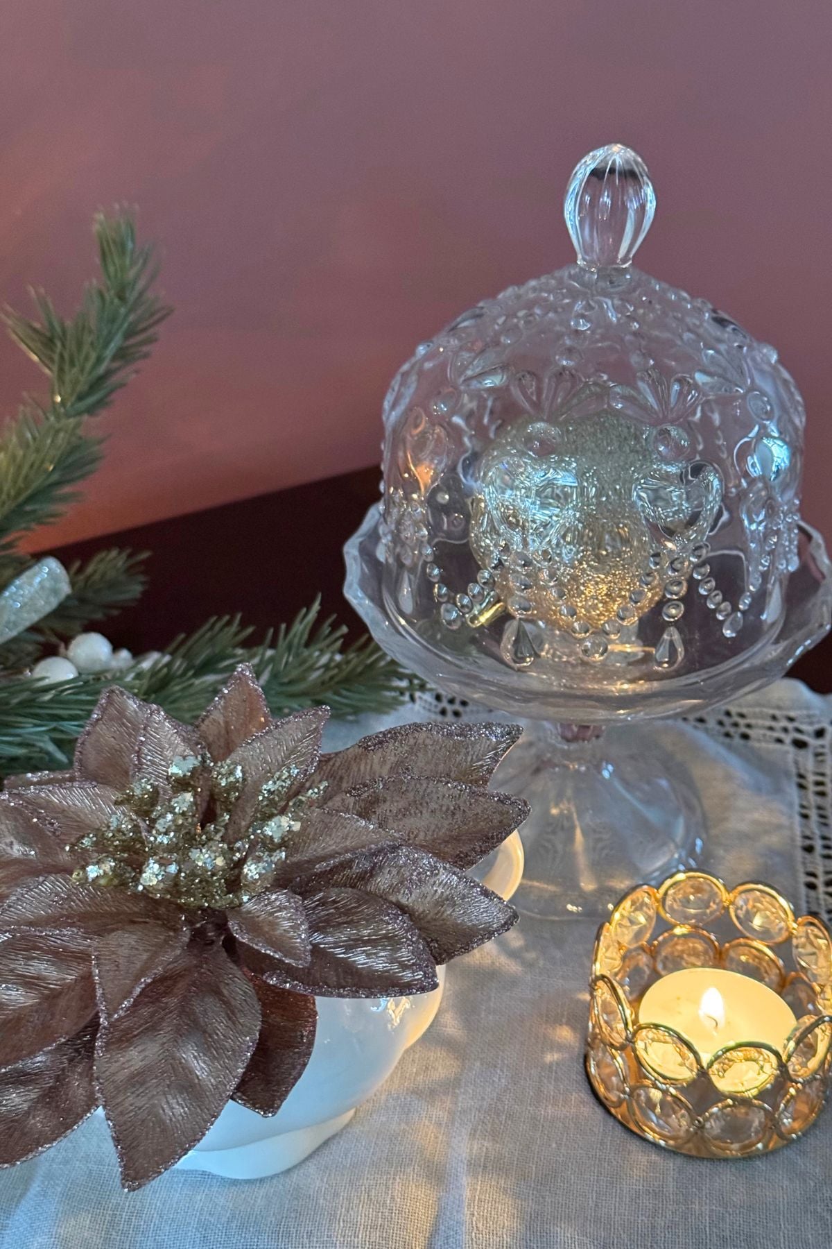 Christmas vignette on sideboard Glass cloche with champagne ornament beside a blush poinsettia and lace doily