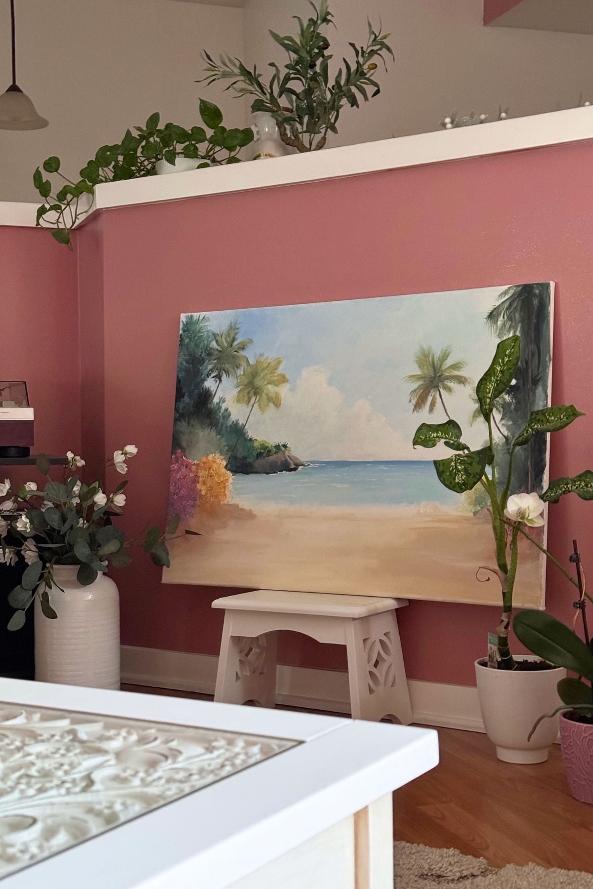 the sentimental art corner in the living room A sentimental art corner in the living room. A beach scene canvas on a white stool surrounded by plants.