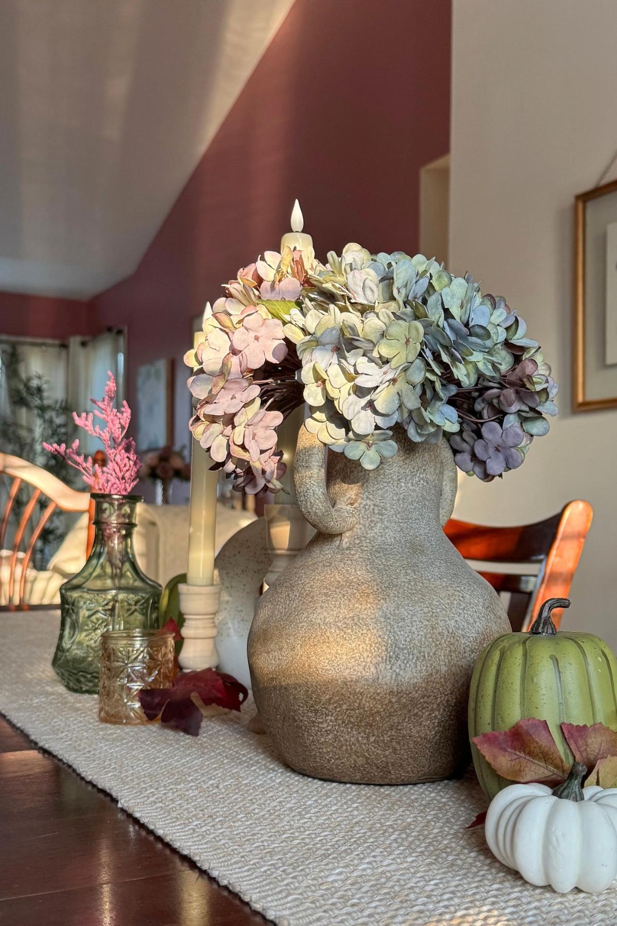 Dining table decor for fall: stone vase, faded hydrangeas, pumpkins, and candle holders.