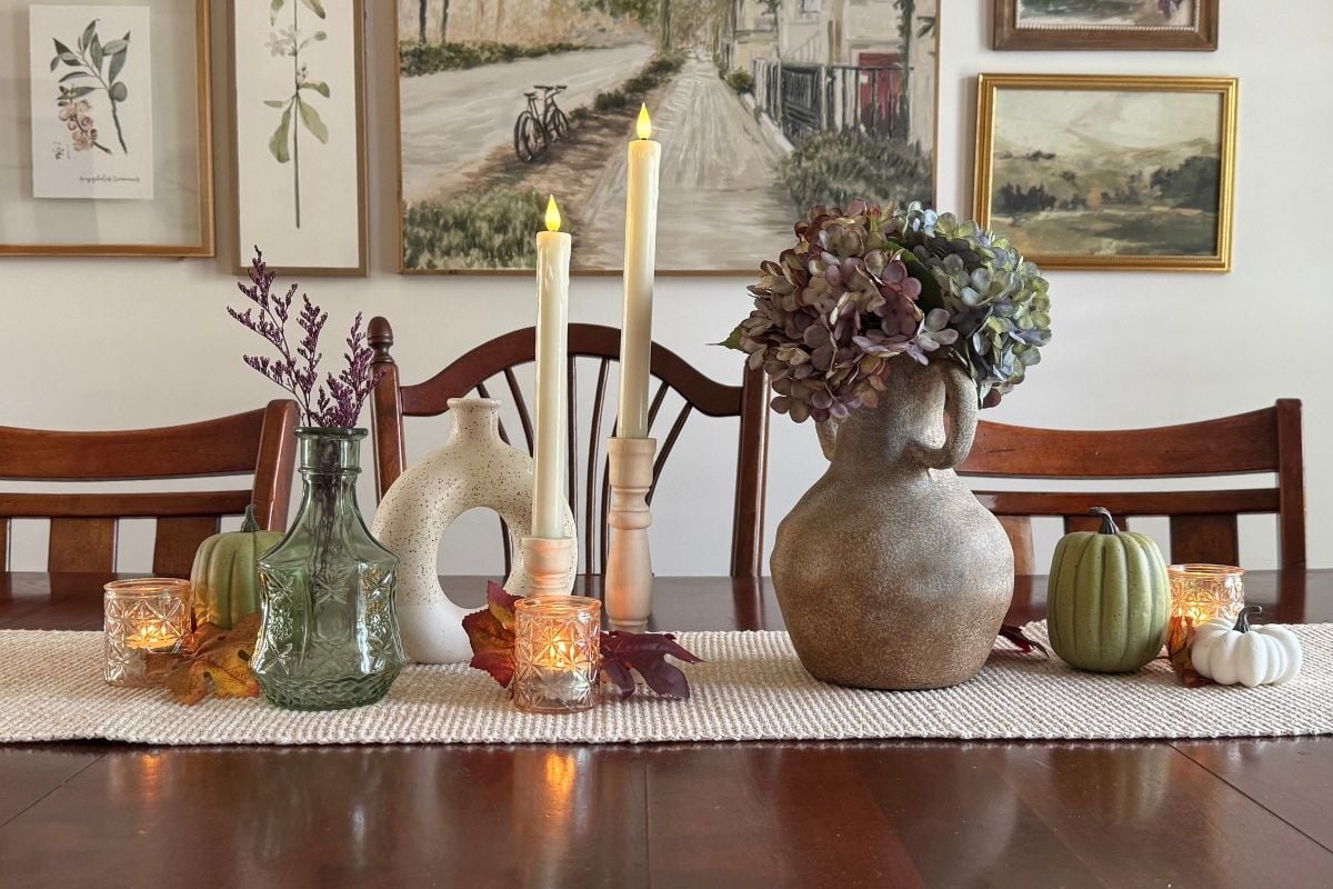 An everyday fall table decor: stone vase, green glass vase, donut vase, candle holders, autumn leaves, sage and cream pumpkins, on a textured table runner on a dark wood dining table.
