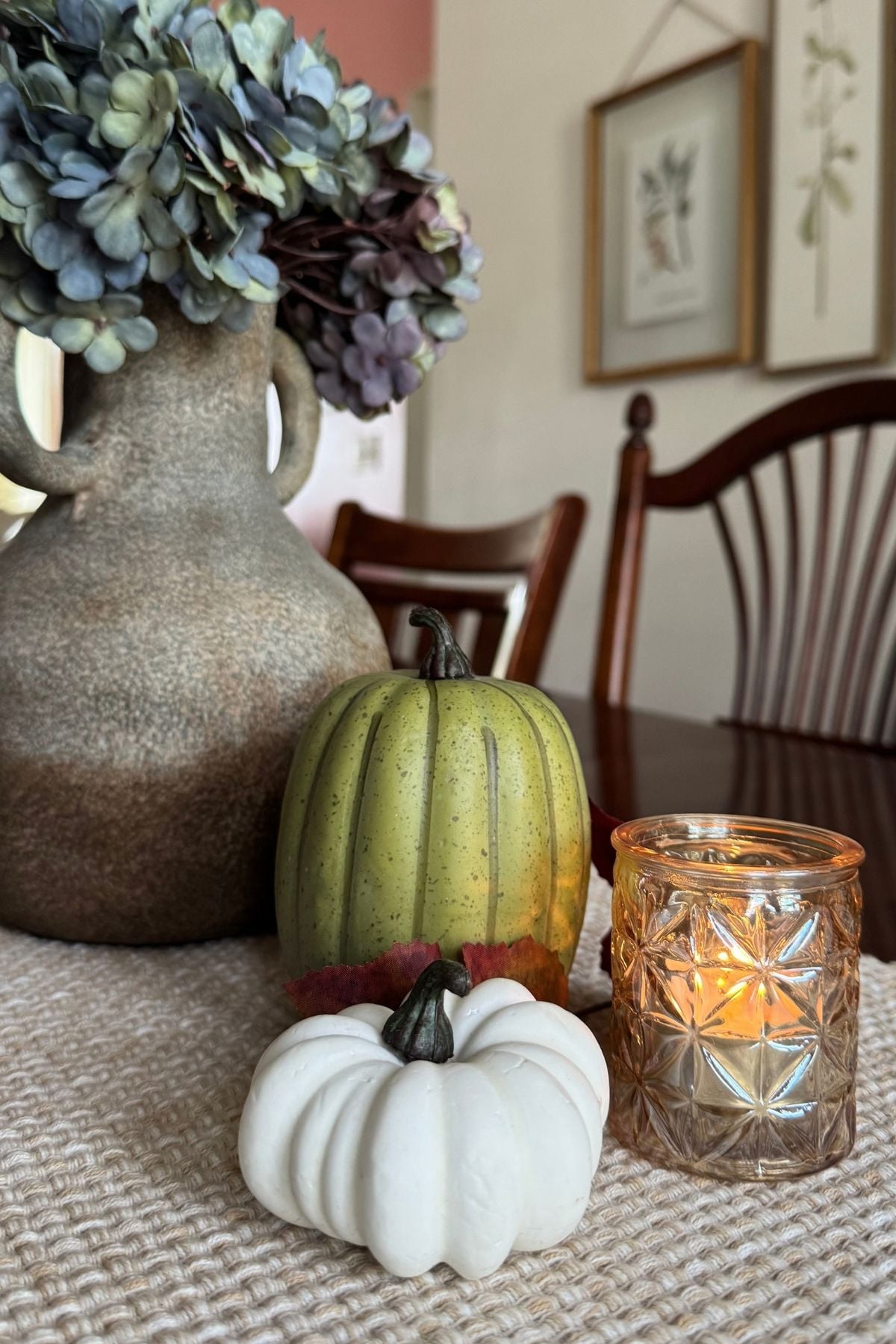An autumn table vignette created with sage and white pumpkins, an amber gold votive candle holder, and a stone vase holding faded hydrangeas.