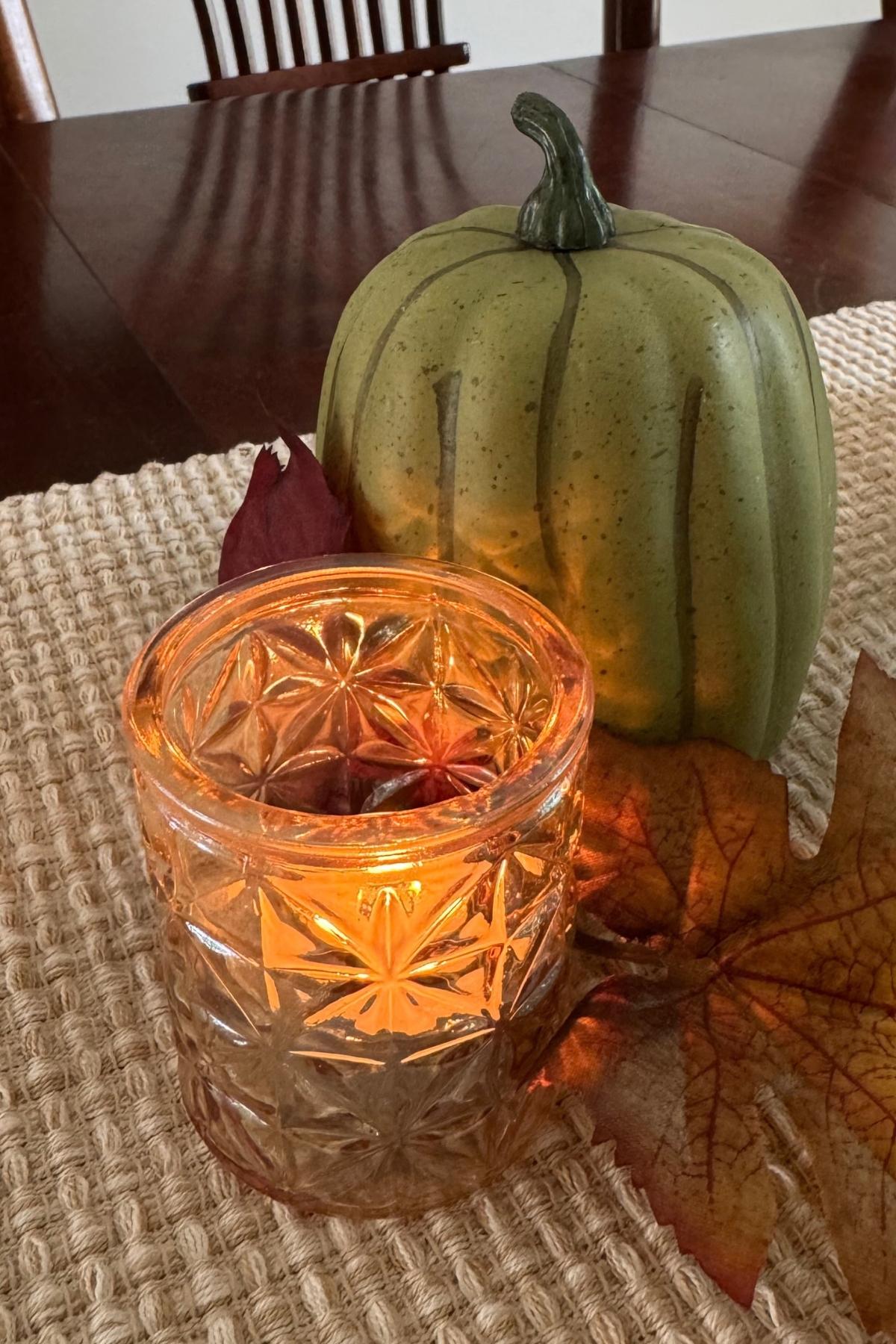 Autumn leaves tucked in between a sage green pumpkin and an amber gold votive candle holder on a woven table runner