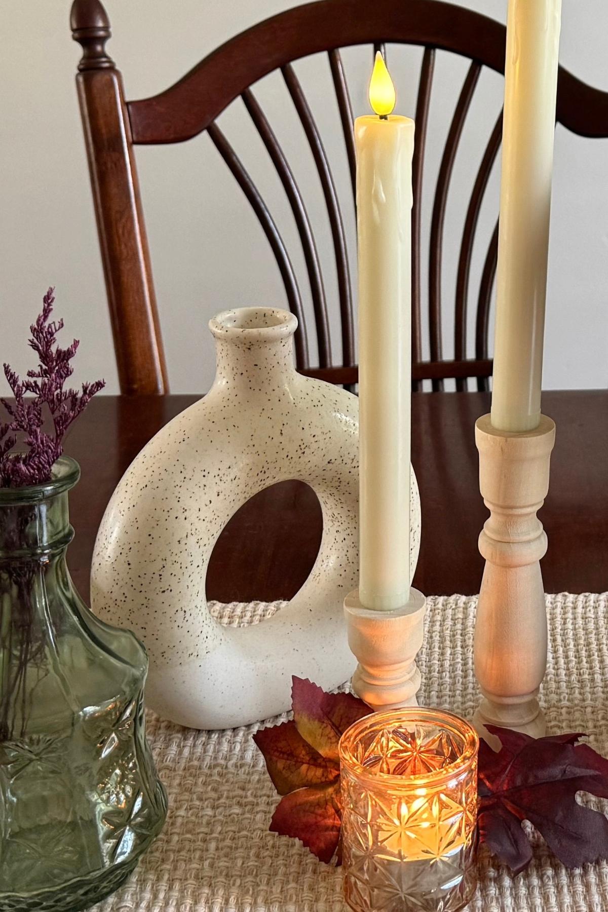 A speckled donut vase, green glass bottle, and flameless candles in wooden candle holders on a fall dining table.